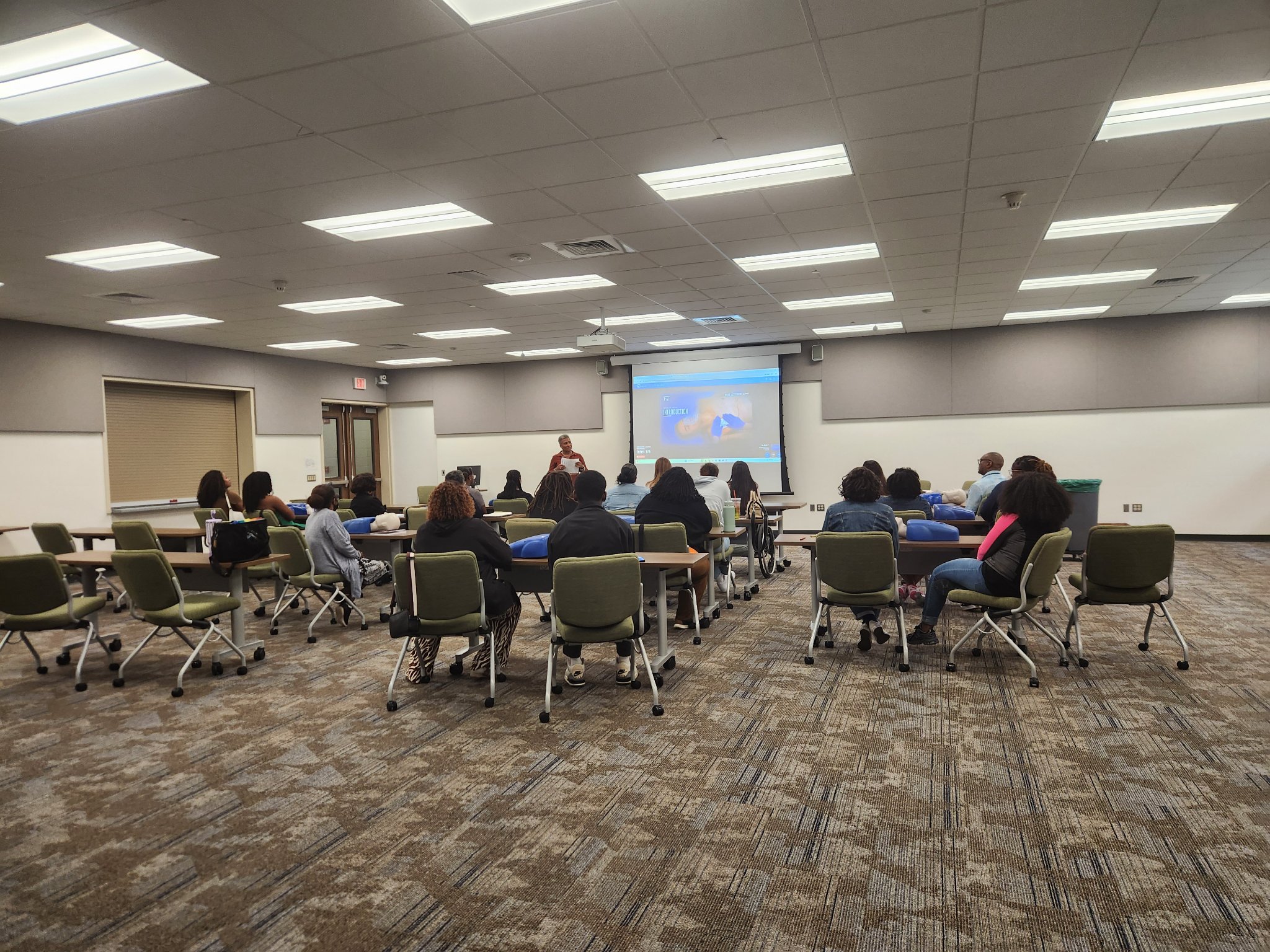 Dantee Johnson teaching a CPR certification class to a room of adult students seated at tables with training mannequins in front of them