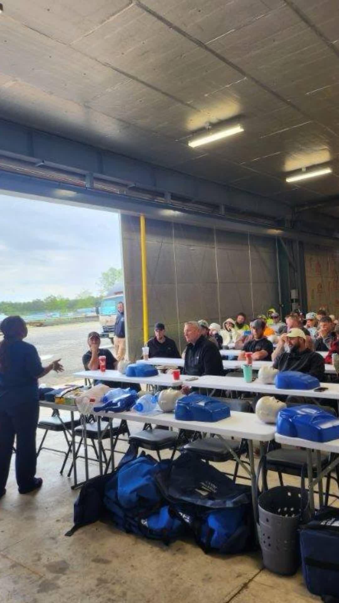 Dantee Johnson instructing a large crew of adult workers seated at tables with CPR mannequins and AED trainers during an on-site workplace class