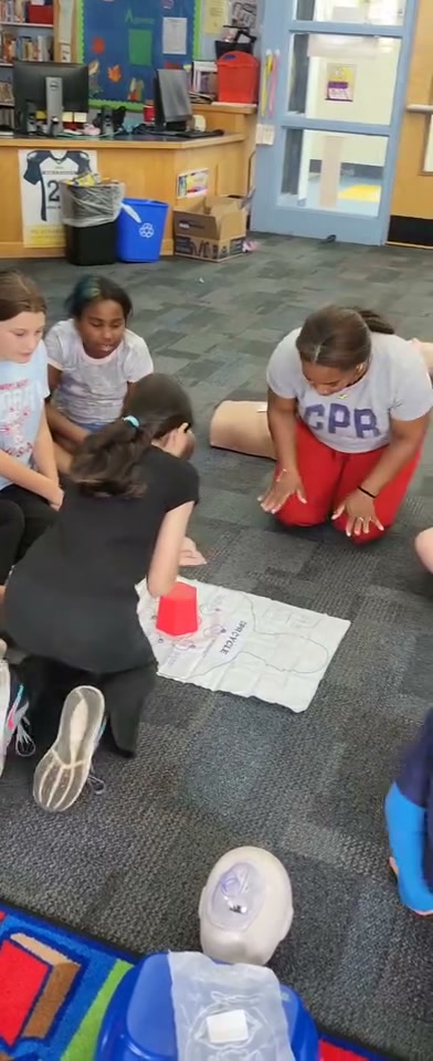 Dantee Johnson in her CPR instructor shirt coaching an elementary student through chest compressions on a practice mat in the school library, classmates watching closely